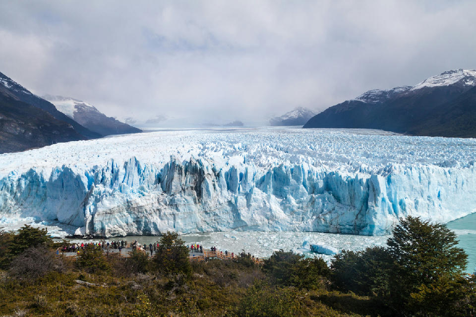 Wide view of Perito Moreno Glacier ice wall with tourists on boardwalks and mountains in the background in Patagonia, Argentina.
