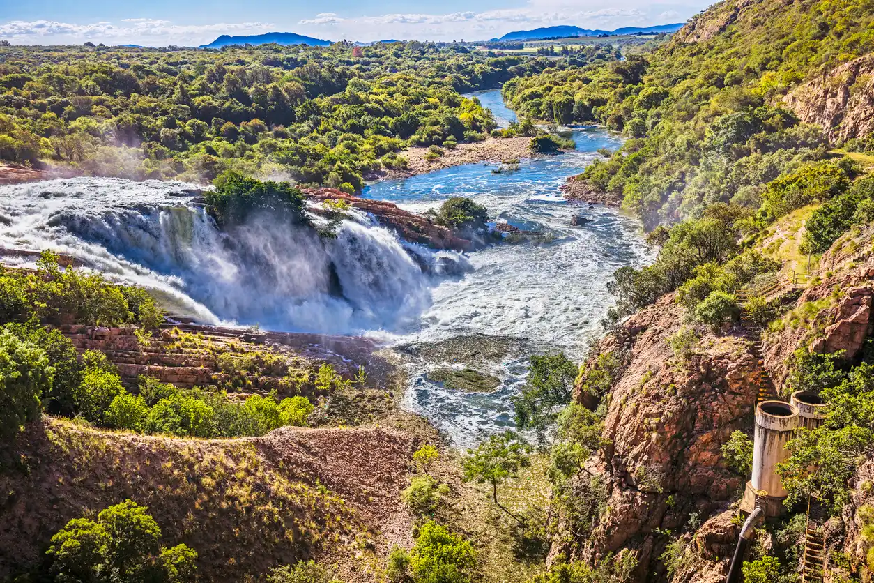 View of the Hartbeespoort Dam spillway and Crocodile River flowing through a rocky gorge below the Magaliesberg, with waterfalls, forested hillsides and surrounding bushveld landscapes.