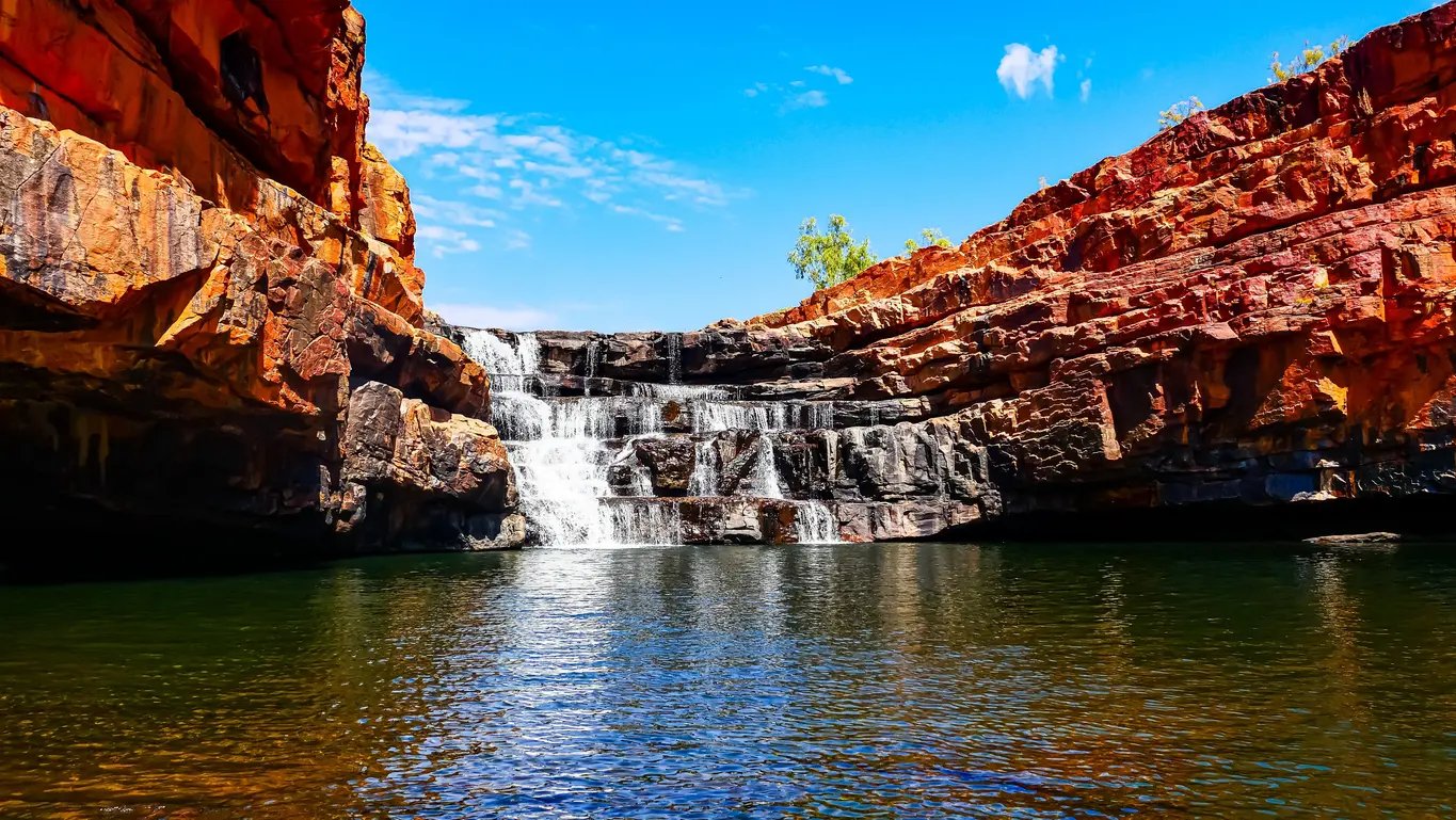 Bell Gorge waterfall cascades over dark rock ledges into a deep pool surrounded by vivid red sandstone cliffs
