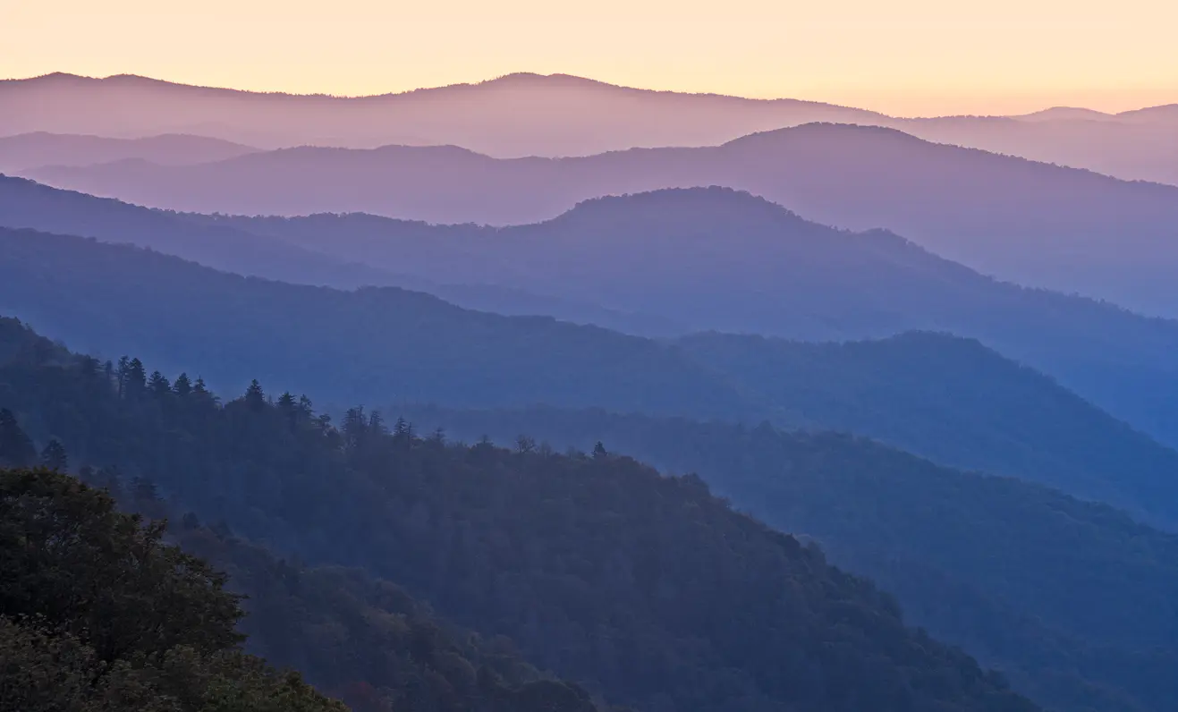 Atmospheric mountain landscape showing overlapping blue ridgelines and forested valleys in the Appalachian and Smoky Mountains at dawn.