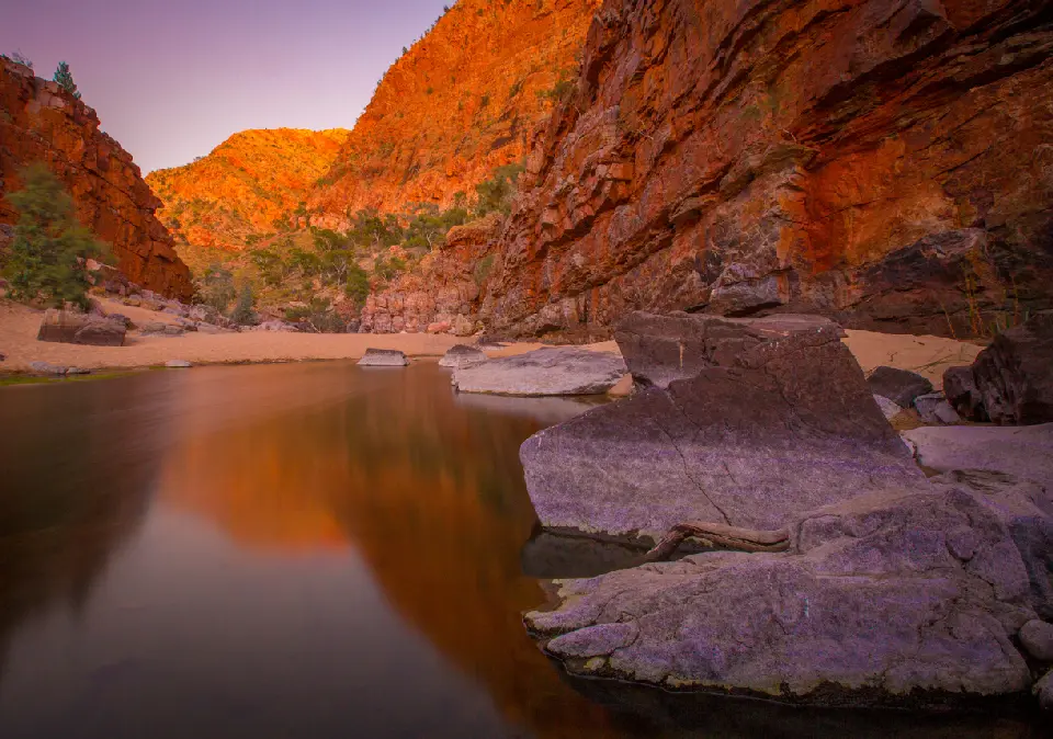 Calm waterhole and large foreground rocks beneath towering red cliffs glowing in warm light at Ormiston Gorge.