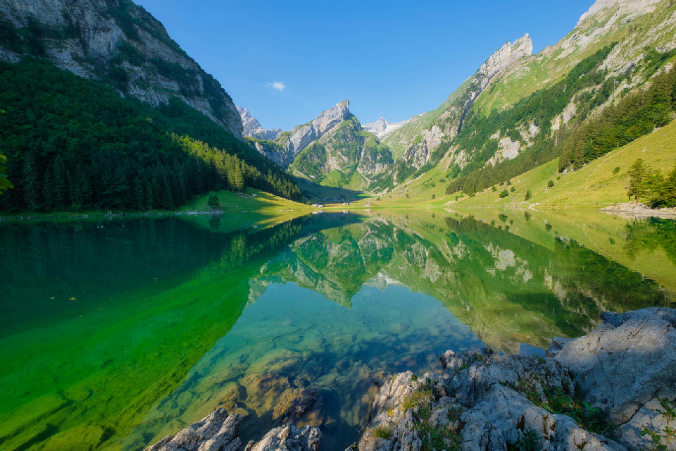Clear alpine lake Seealpsee reflecting surrounding mountains and green slopes in the Alpstein region of Switzerland, with rocky shoreline in the foreground.