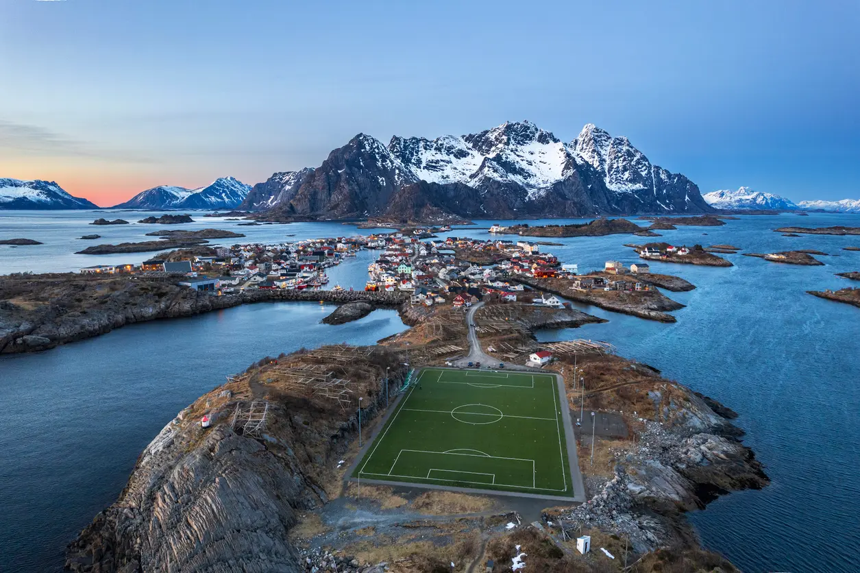 Aerial view of Henningsvær on Austvågøya in the Lofoten Islands, showing the famous football field on a rocky island surrounded by fjords and snow-covered mountains at dusk in Nordland, Norway.
