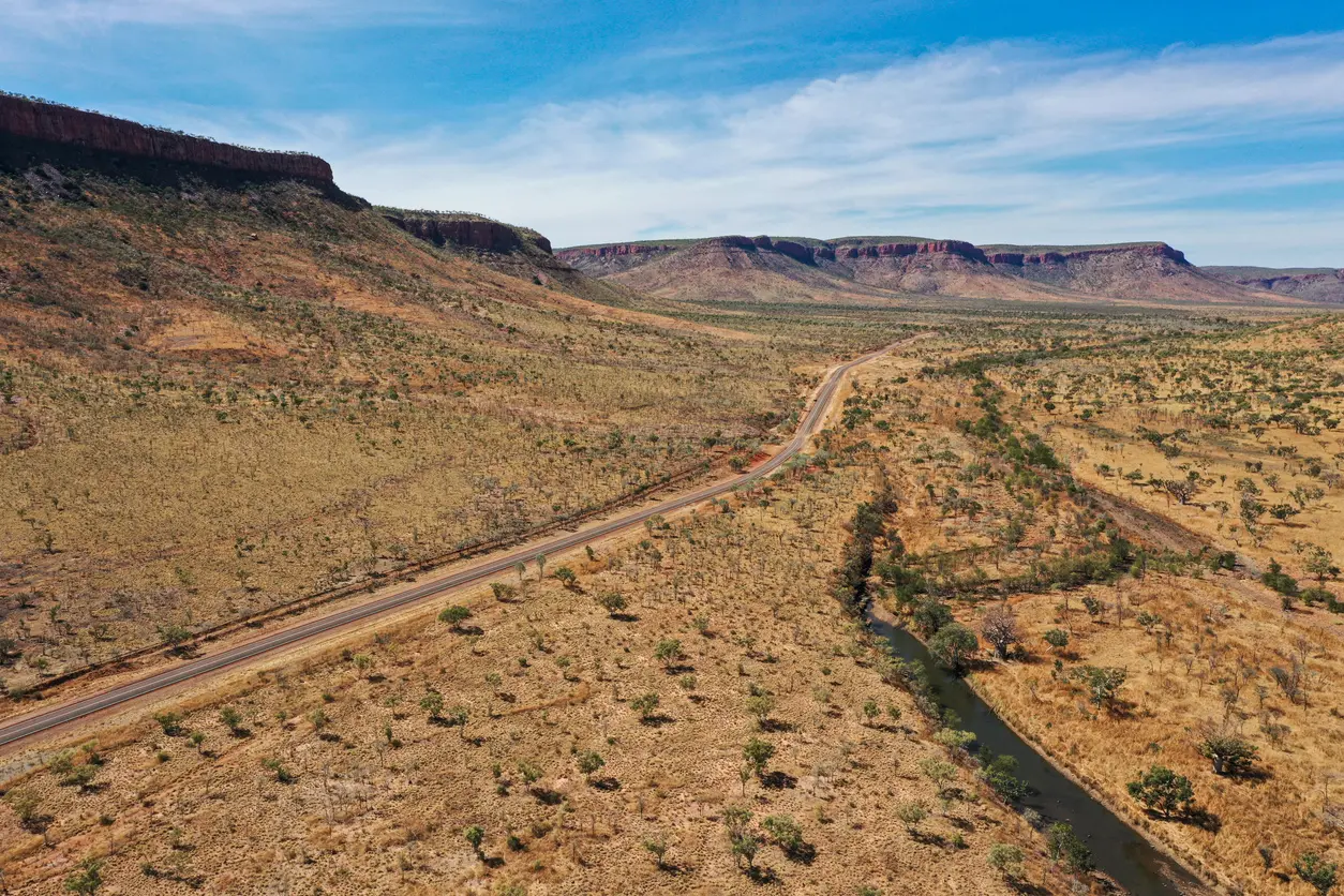 Aerial view of a remote road and river winding beneath rugged sandstone escarpments across the East Kimberley landscape.