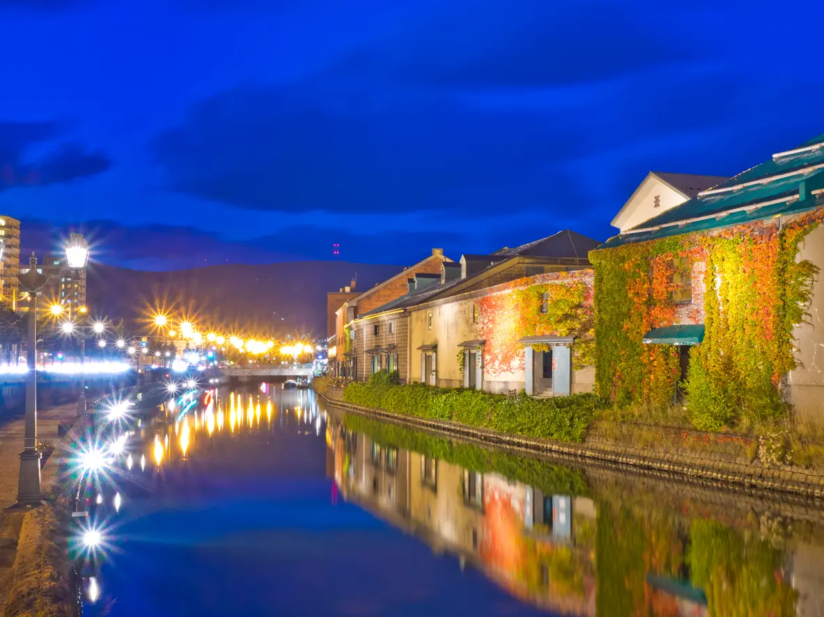 Historic warehouse buildings along Otaru Canal reflected in calm water at dusk, with street lamps illuminating the scene in Hokkaido, Japan.