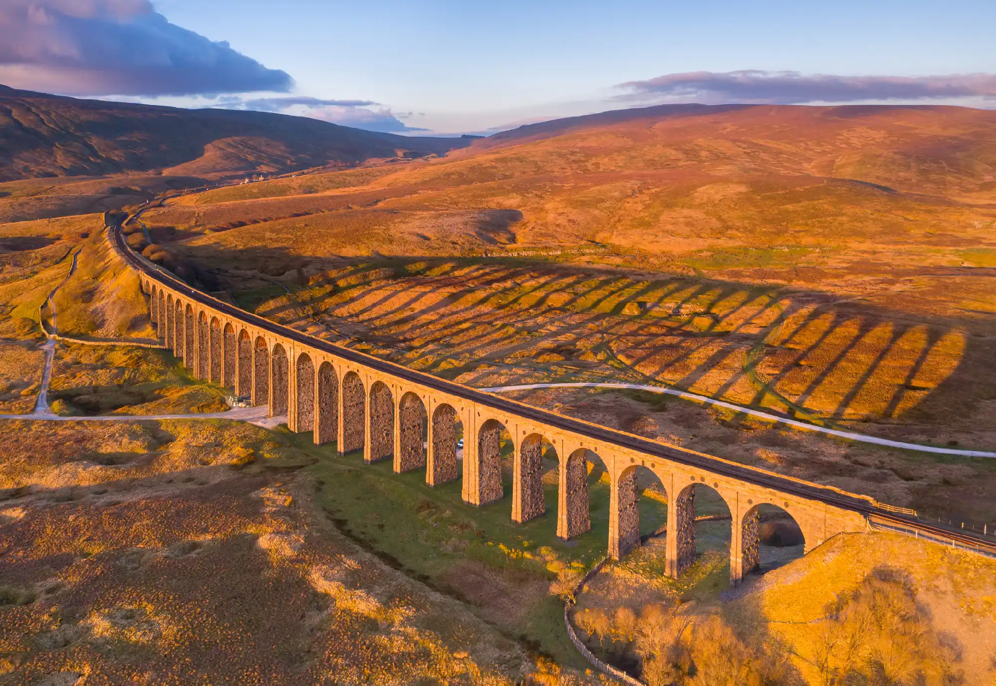 Elevated view of Ribblehead Viaduct illuminated by golden evening sunlight, with long shadows from its stone arches stretching across Batty Moss in the Yorkshire Dales.