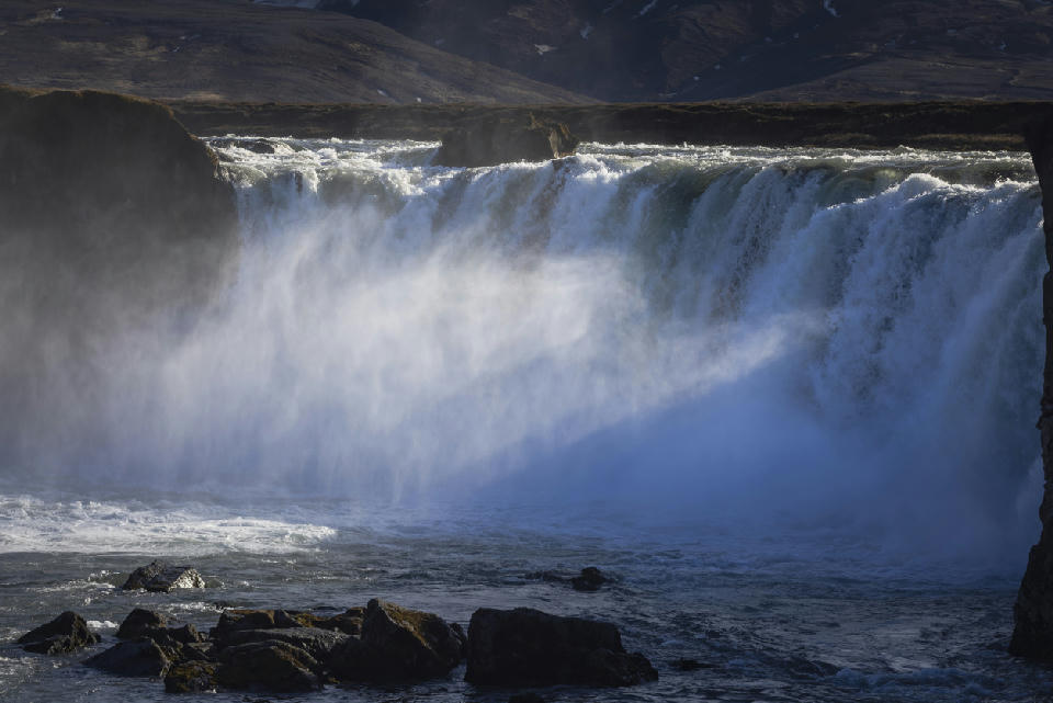 Goðafoss waterfall in the Mývatn region of North Iceland, showing powerful water flowing over a broad curved cliff into a rocky river below.