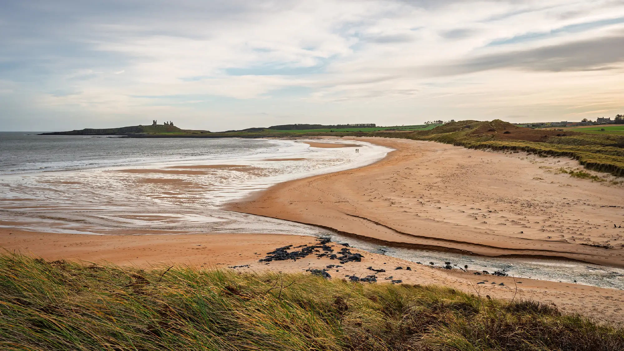 Elevated view of Embleton Bay in Northumberland, showing its sweeping sandy beach, Embleton Burn, grassy dunes and Dunstanburgh Castle on the distant headland.