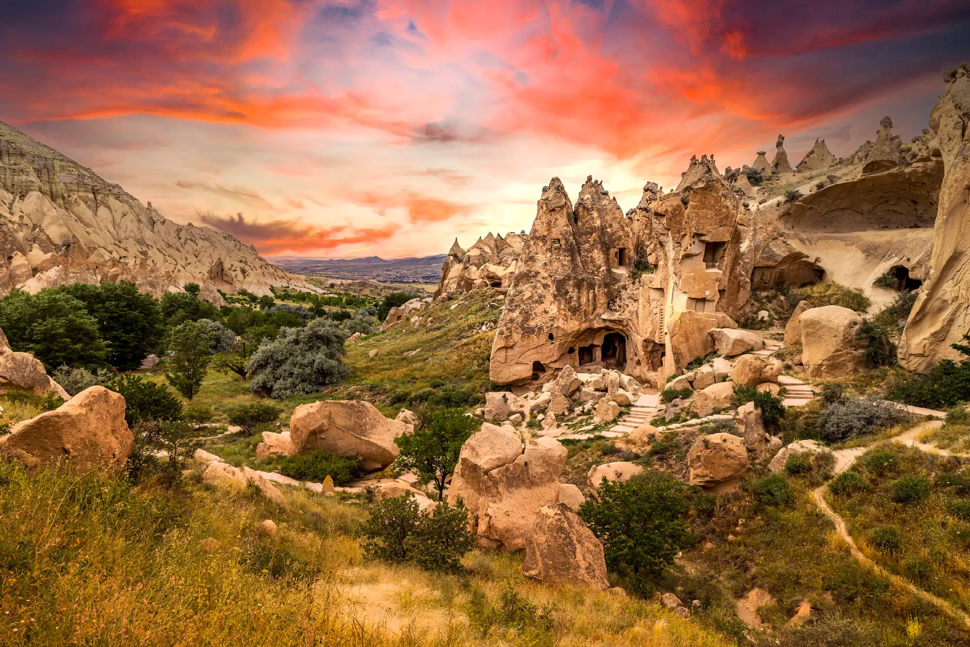Ancient cave houses carved into volcanic rock at Zelve Open Air Museum in Cappadocia, Turkey, surrounded by dramatic valleys and fairy chimney formations beneath a colourful sunset sky.