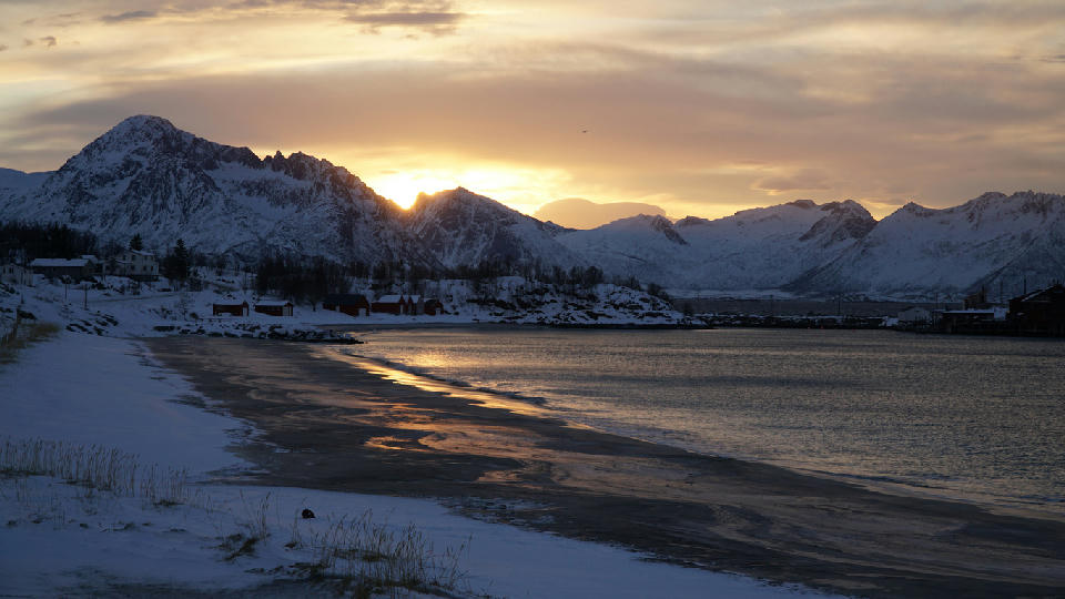 Curving snowy beach at Bøvær with wet sand reflections, red cabins, and snow-covered mountains at sunset on Senja island, Norway