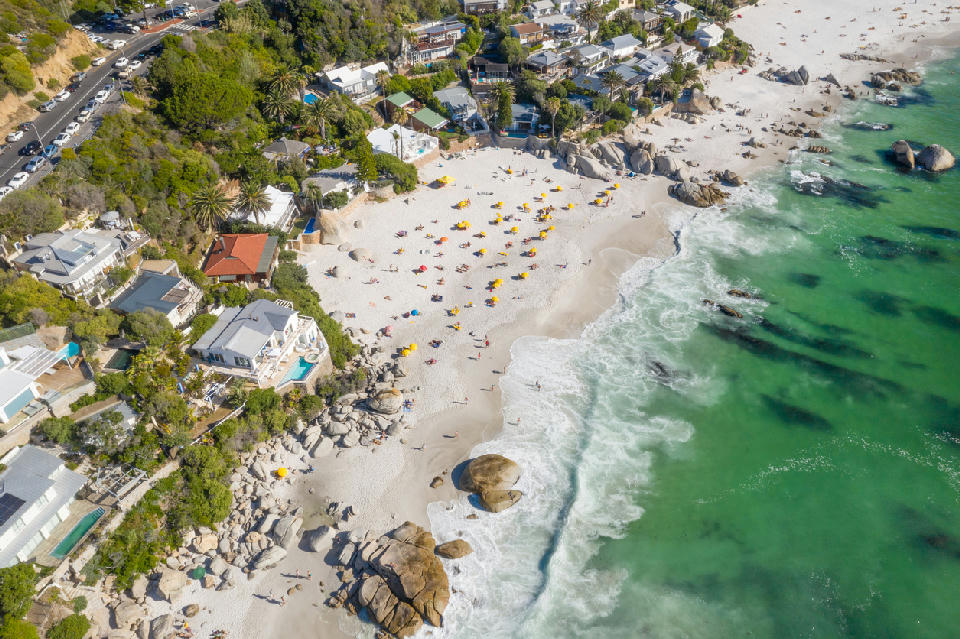 Drone photograph of Clifton Beach in Cape Town showing beachgoers on white sand, large granite rocks along the shoreline and clear green Atlantic waves beside hillside homes.