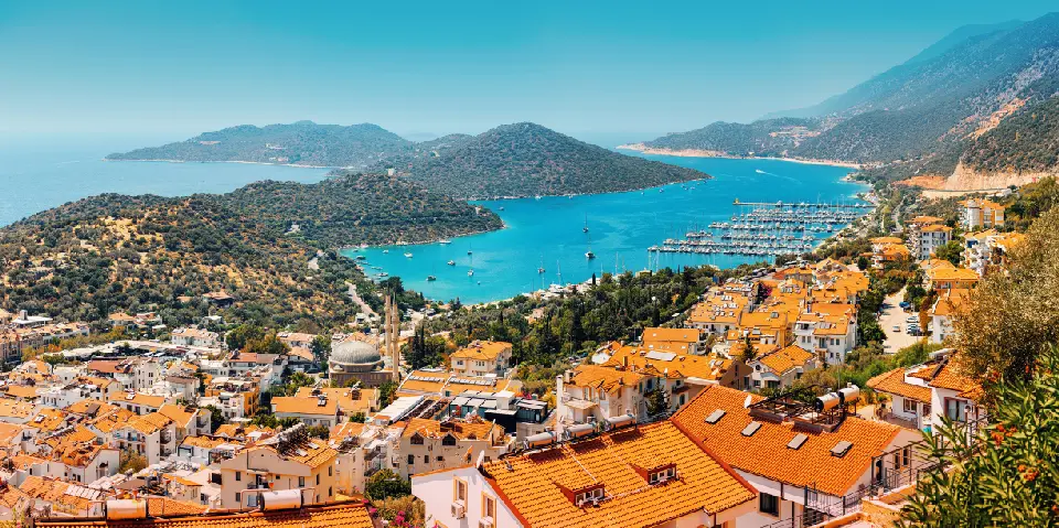 Panoramic view over Kaş, with orange-tiled rooftops, a marina filled with yachts, turquoise bays and the mountainous Mediterranean coastline.