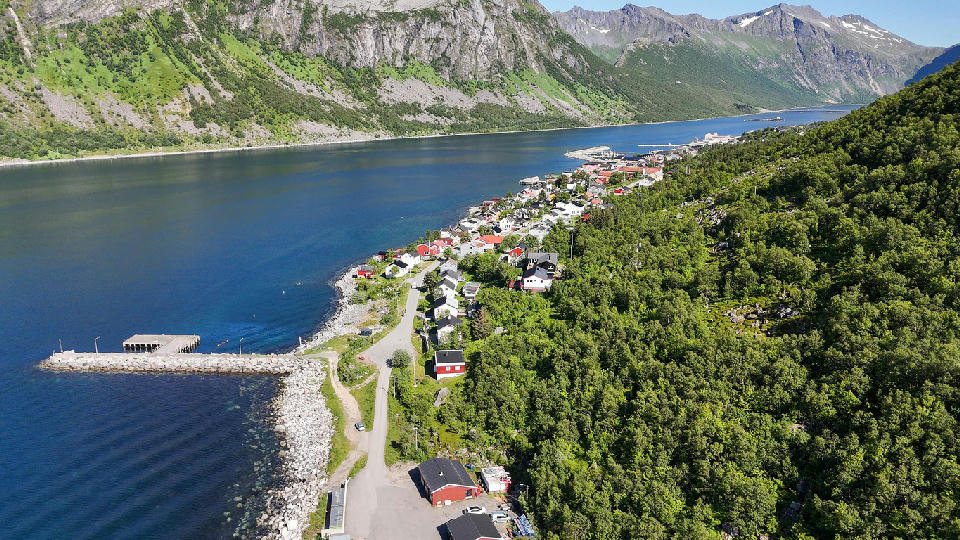 Aerial view of Gryllefjord village on Senja with houses along a curved shoreline, harbour pier, and steep green mountains above a fjord