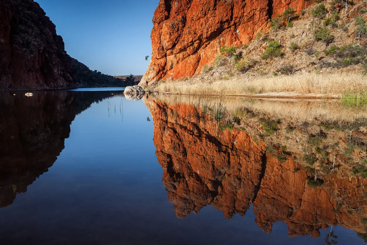 Red-orange rock formations reflect in the calm, glassy water of the Glen Helen Gorge waterhole beneath a clear blue sky.