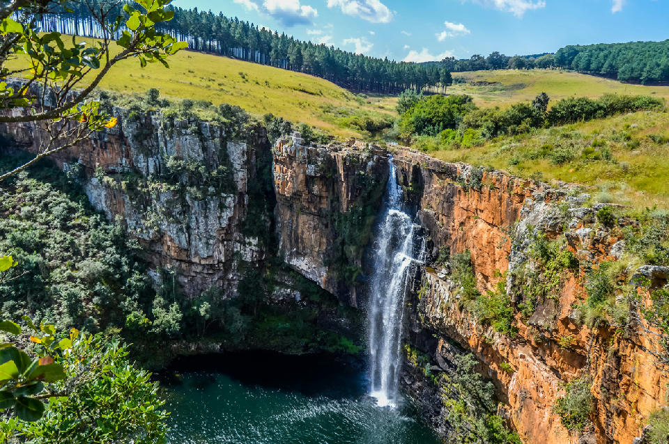 Berlin Falls dropping over orange rock cliffs into a deep plunge pool surrounded by grassland and forest in Mpumalanga.