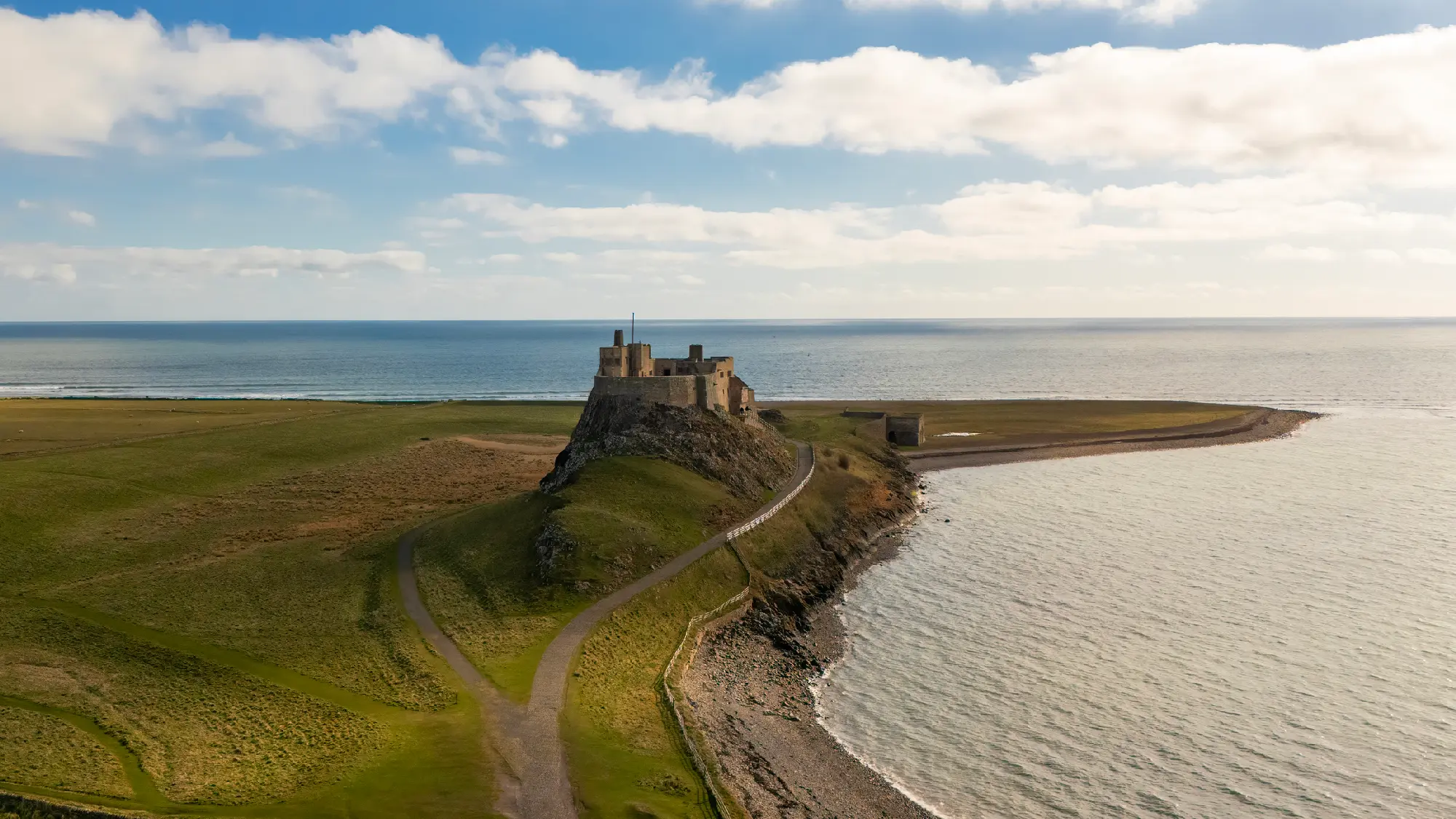 Aerial view of Lindisfarne Castle on a rocky hill beside the sea on Holy Island, Northumberland, showing coastal paths, grassy tidal land and the surrounding shoreline.