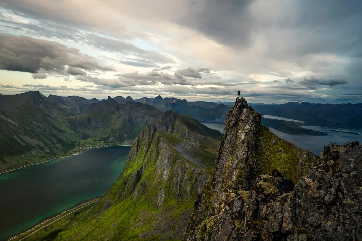 Hiker standing on rocky ridge at Hesten viewpoint Senja Norway overlooking Segla peak and Mefjorden fjord with steep cliffs and layered mountains.