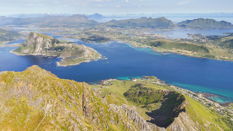 Aerial view from Stornappstinden toward Offersøykammen on Vestvågøya in the Lofoten Islands, showing fjords, green farmland and mountain ridges along the Norwegian coast.
