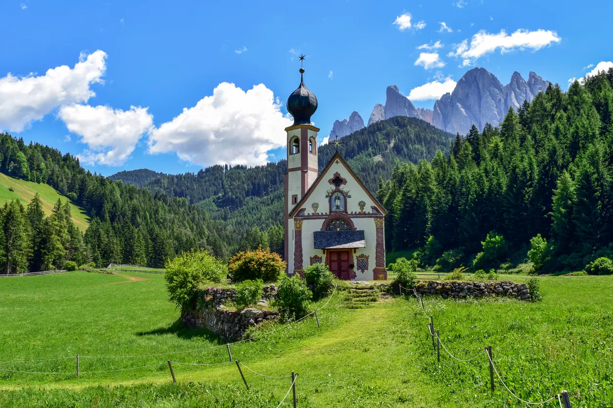 St. Johann Church on a grassy hillside in Val di Funes, with open meadows in the foreground and the Odle mountain group rising behind.