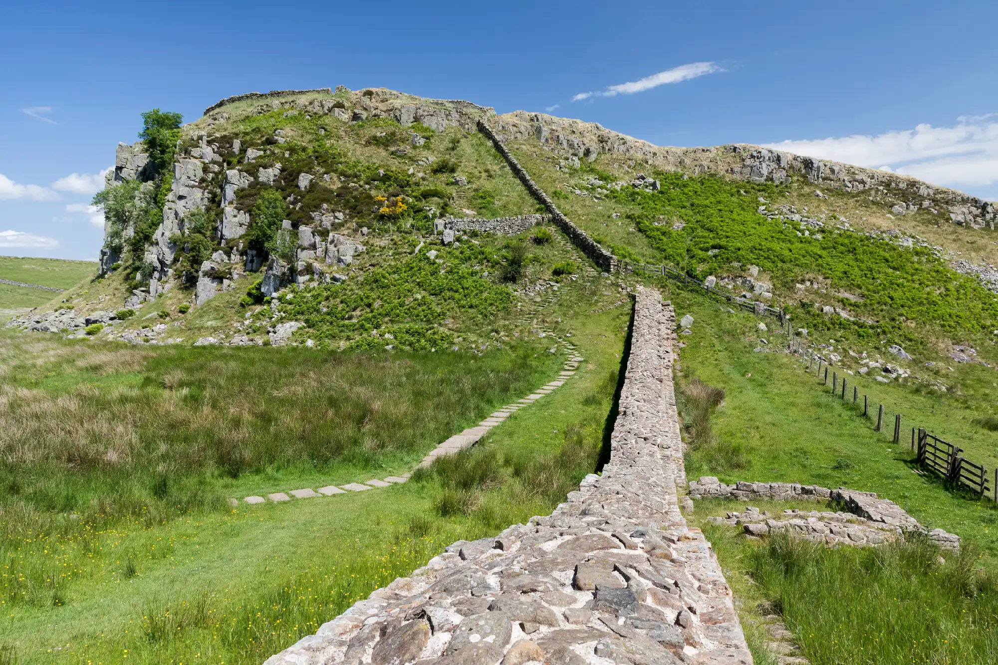 Hadrian's Wall ascending a steep grassy and rocky hillside at Peel Crags near Steel Rigg, Northumberland, England, beneath a blue sky.