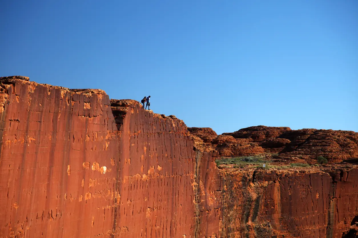 Hikers cross the rim above the immense red sandstone walls of Kings Canyon beneath a clear blue sky.