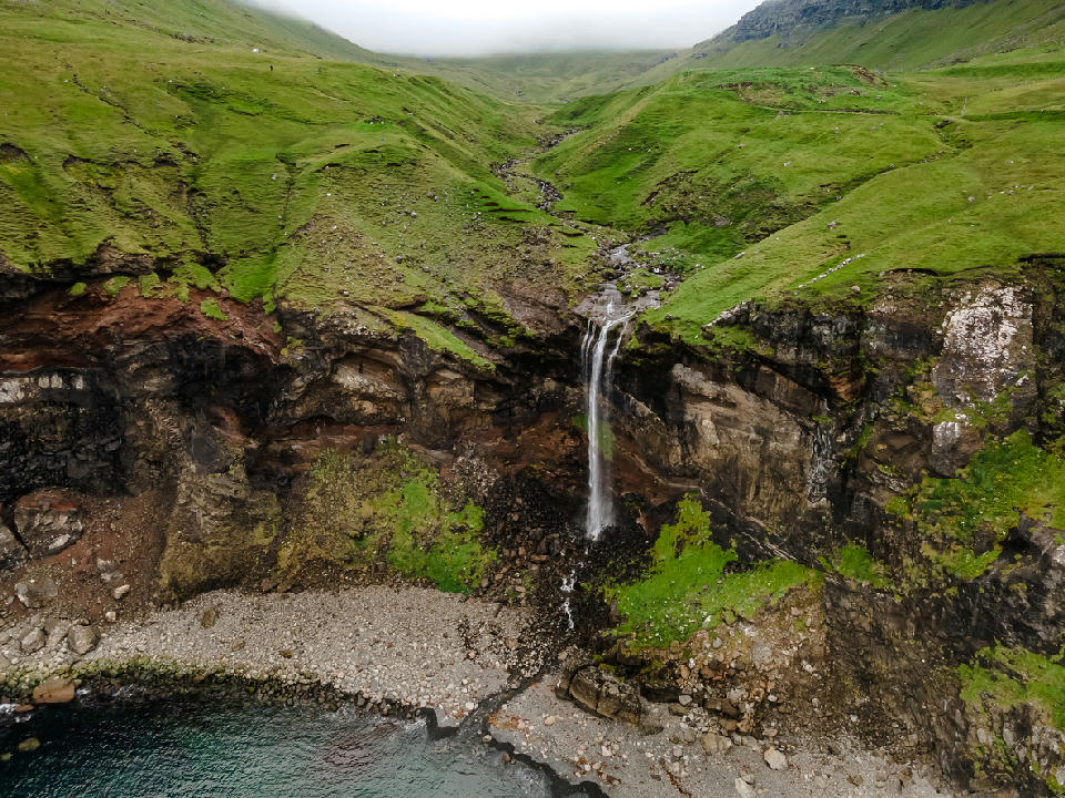 Aerial view of Skarðsáfossur waterfall flowing over basalt cliffs into the Atlantic Ocean on Vágar Island, Faroe Islands.