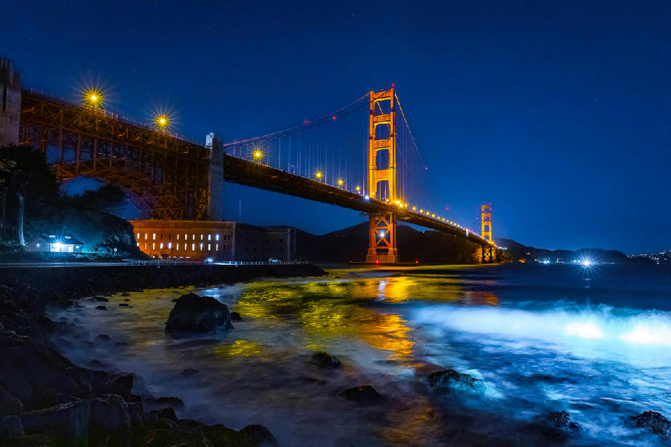 Golden Gate Bridge illuminated at night with reflections in the bay and rocks in the foreground