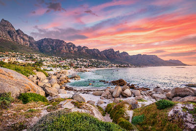 Camps Bay coastline with boulder-lined foreground, white sand beach, and the Twelve Apostles mountain range at sunset, Cape Town.