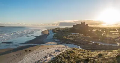 Four racehorses return from their early-morning exercise along a snow-covered Bamburgh Beach, with Bamburgh Castle silhouetted against the golden winter sunrise.