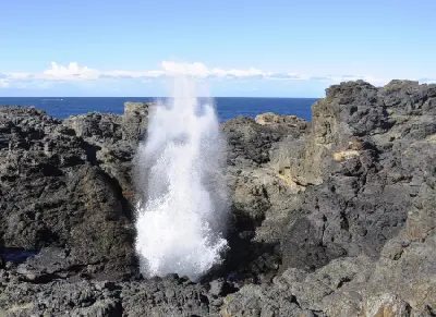 Powerful plume of seawater erupts from the Kiama Blowhole between dark coastal rock formations beside the Pacific Ocean.