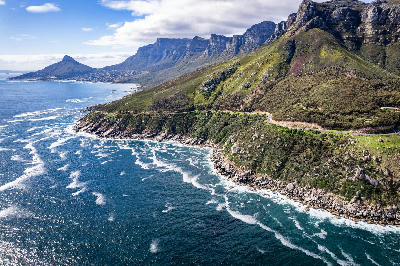 Aerial view of Victoria Road winding along a rocky Atlantic coastline beneath the Twelve Apostles mountains in Cape Town.