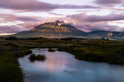 Ol Doinyo Lengai volcano illuminated by early morning light above a reflective stream and Maasai plains near Lake Natron, Tanzania.