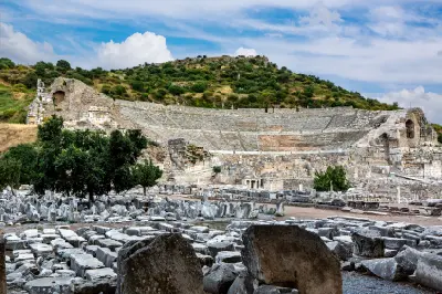 Wide view of the Great Theatre of Ephesus, with its semicircular stone seating rising behind scattered marble ruins and trees.