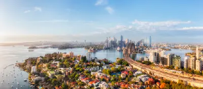 Wide aerial view over Kirribilli towards Sydney Harbour, with the Opera House, Harbour Bridge and CBD skyline in warm late-afternoon light.