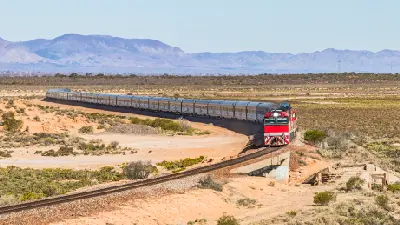 Long passenger train with red locomotive travelling through the South Australian outback, with dry scrubland and the Flinders Ranges mountains in the background.