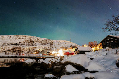 Northern lights faintly visible above snowy coastal village in Ersfjord with illuminated houses at night