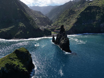 Volcanic sea stacks rising from the Atlantic at Ilhéus da Ribeira da Janela, framed by deep blue water and steep, terraced green cliffs along Madeira’s north coast.