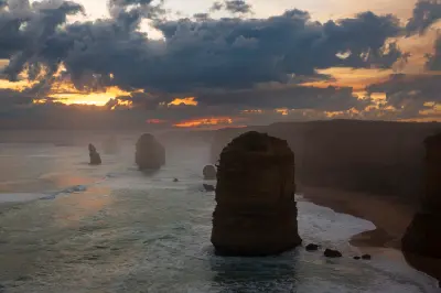 Twelve Apostles sea stacks beneath dramatic clouds and coastal mist at sunset, Victoria, Australia.