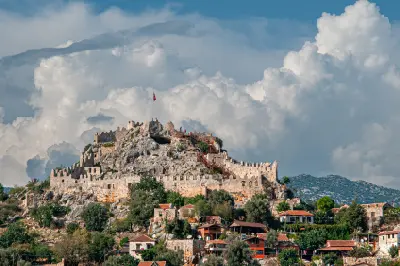 Simena Castle in Kaleköy, Kekova, Turkey, standing on a rocky hill above traditional stone houses and red-tiled roofs beneath dramatic clouds.