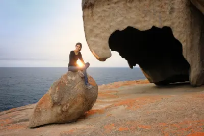 Woman sitting on a granite boulder beside a large overhanging rock formation at Remarkable Rocks, overlooking the Southern Ocean on Kangaroo Island, South Australia.