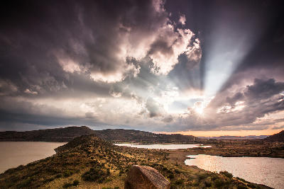 Dramatic sunset and storm clouds over Gariep Dam, with sun rays shining across the reservoir and surrounding semi-arid landscape.