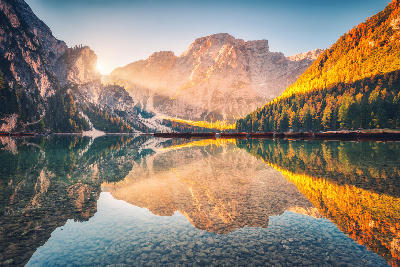 Autumn sunrise at Lago di Braies, with golden light on surrounding Dolomite peaks, calm water reflections, and trees with orange leaves in the Italian Alps.