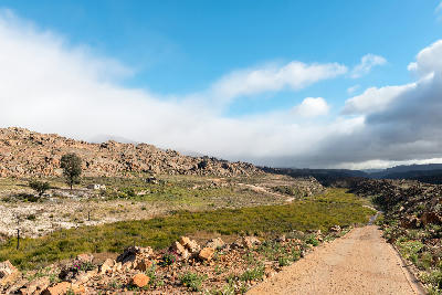 Gravel road winding through rocky mountain terrain between Matjiesrivier and Wupperthal, with a remote farmhouse and historic ruins visible in the landscape.