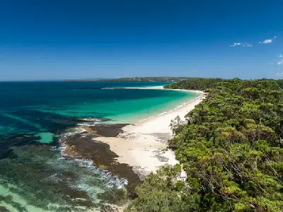 White sand curves around Huskisson Beach beside turquoise water, rocky reefs and dense coastal forest on Jervis Bay.