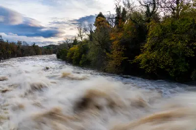 Fast-flowing water rushing over Aysgarth Falls, framed by autumn woodland beneath a cloudy sky in the Yorkshire Dales, England.