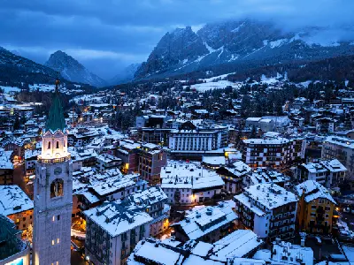 Aerial view of Cortina d’Ampezzo illuminated at night, with a snow-covered mountain village and bell tower in the foreground in the Dolomites, Veneto, Italy.