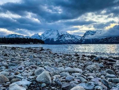 Rocky shoreline leading to Jackson Lake with snow-covered Teton Range under dramatic clouds in Grand Teton National Park, Wyoming, USA.