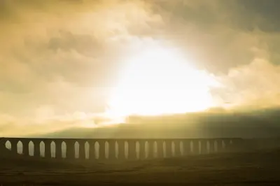 Backlit Ribblehead Viaduct at sunset, with its repeating stone arches silhouetted against golden clouds above the dark moorland.