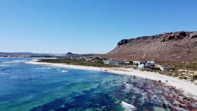 Drone view of Elands Bay on South Africa's West Coast, showing turquoise ocean water, sandy shoreline, coastal houses and the rocky escarpment rising behind the bay.