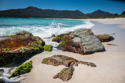 Granite boulders beside turquoise waves and white sand at the northern end of Wineglass Bay, with rugged mountains rising behind in Freycinet National Park, Tasmania.